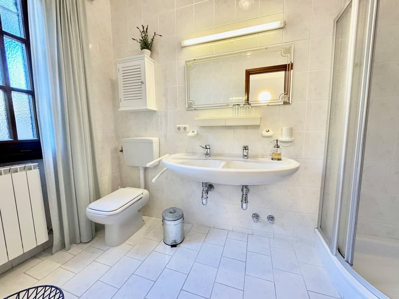 Spacious bathroom with white tile flooring, double sink vanity, toilet, and a storage cabinet. The bathroom has a large window and appears to be well-lit.