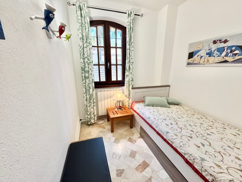 Bedroom with wooden framed windows, patterned curtains, wooden side table with lamp, tiled floor, and a bed with patterned bedding.
