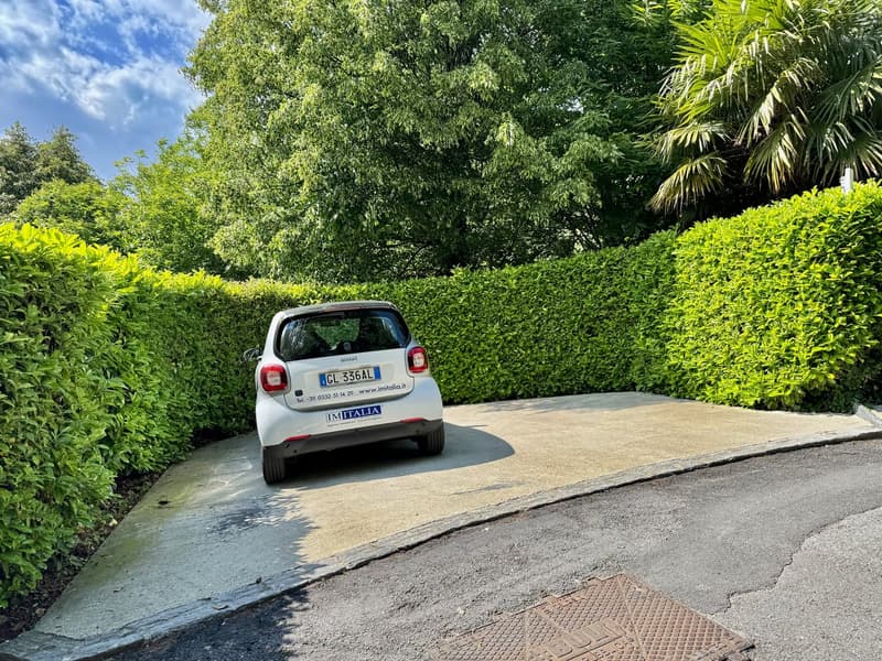 Small car parked in a parking area with a concrete surface, surrounded by well-maintained hedges.