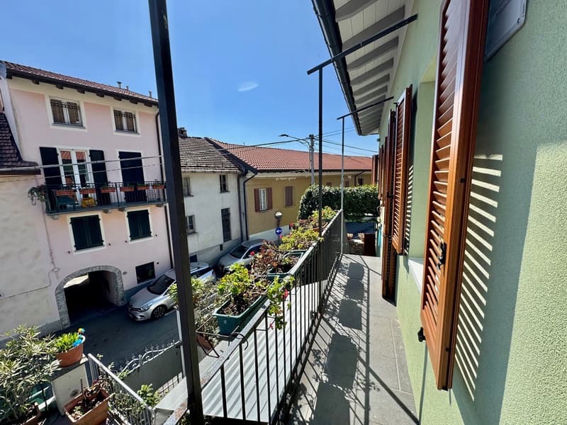 The image shows a multi-story apartment building with a balcony or terrace visible. The building has a mix of pink and yellow facades, with shuttered windows and a tiled roof. There are cars parked on the street below, and some potted plants and greenery