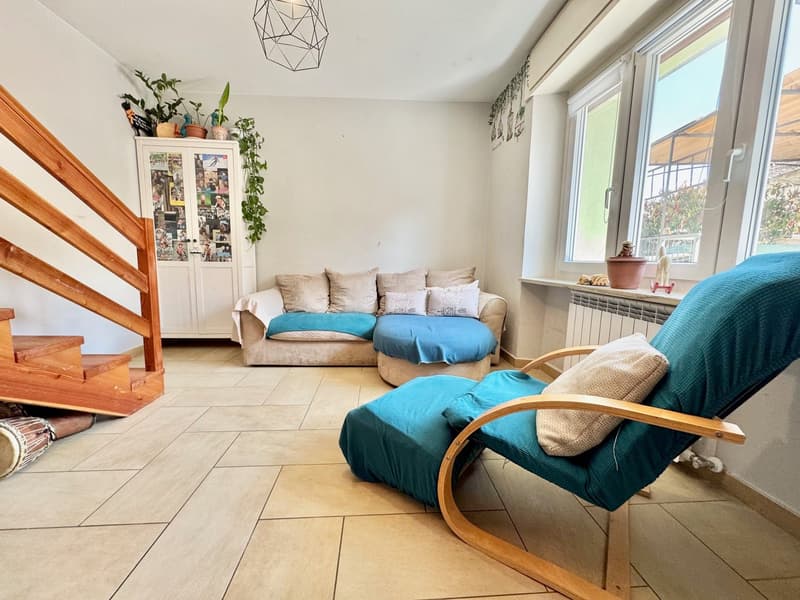 Cozy living room with a large sofa, wooden rocking chair, and potted plants. The room has large windows that provide natural light and a view of the outdoors. The floor is tiled and the room has a modern, minimalist decor.