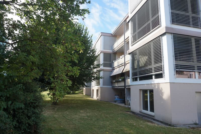 Modern apartment building with beige walls, windows, and balconies, greenery and trees in the background.