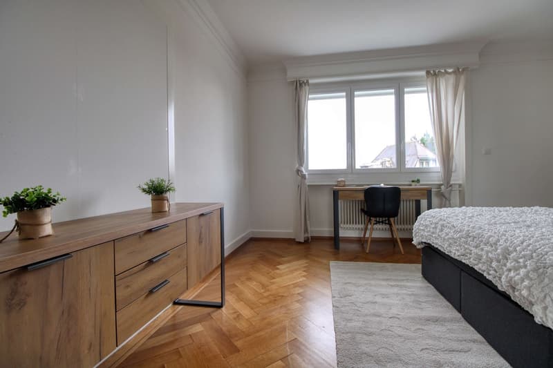 modern bedroom with a bed, desk, chair, white carpet, wooden parquet floor, side table with potted plants, large window with curtains