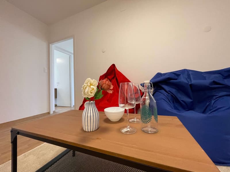 A living room with a wooden table, a vase with flowers, wine glasses, a bowl, and a bottle. There is a blue bean bag chair and a red bean bag chair in the room. The room has a white wall and a wooden floor. There is a door leading to another room.
