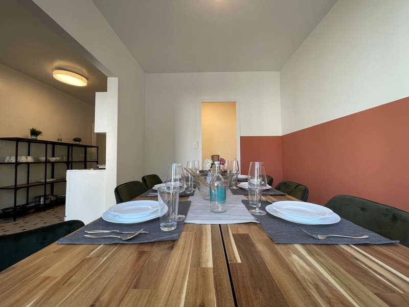 Dining room with a long wooden table, set for multiple people, white plates, glasses, and cutlery, green chairs, shelves with plants and kitchenware, white and orange walls, ceiling light
