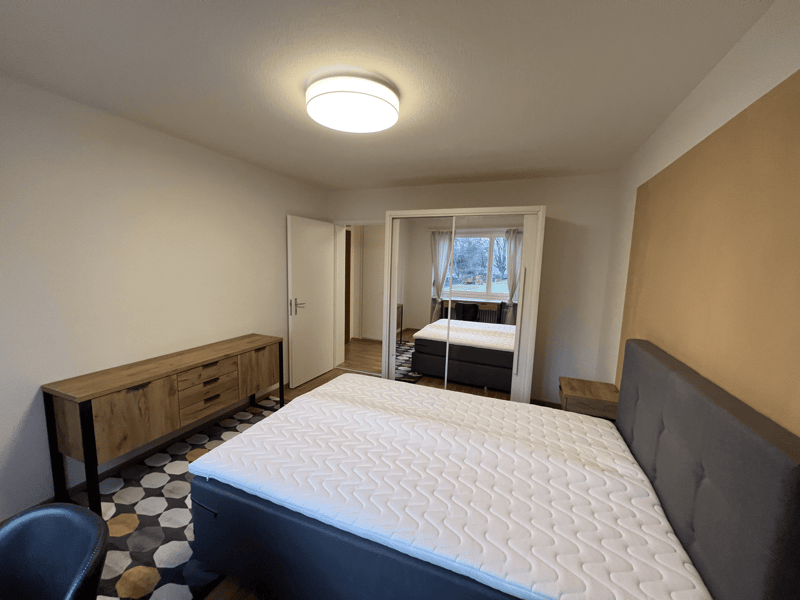 Bedroom with a double bed, wooden cabinet, and a chair. The room has a carpet, a ceiling lamp, and a mirror. There is a window with curtains and a view of the outside.