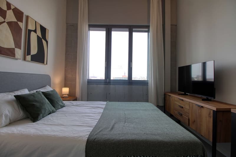 A bedroom with a bed, nightstand, lamp, TV, and window with curtains.