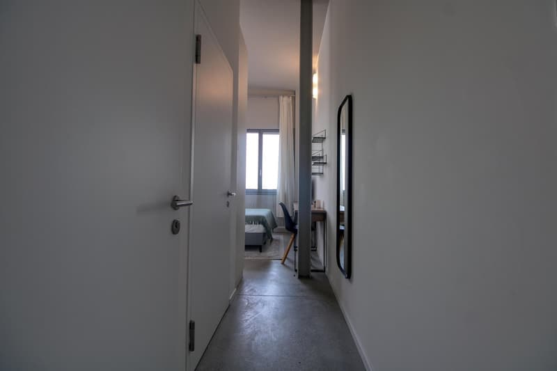 Empty hallway with white walls, concrete floor, a door, a mirror, a desk, and a chair.