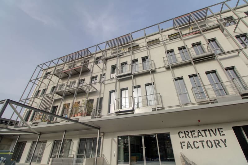 Modern apartment building with multiple balconies, glass doors, and windows, metal railings, and a sign reading 'Creative Factory'.