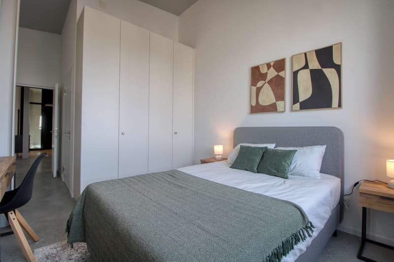 Bedroom with bed, nightstands, lamps, and artwork on the wall. There is a rug on the floor and a chair near the desk.