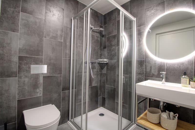 modern bathroom with gray tiles, glass shower enclosure, white toilet, white sink, round mirror, wooden shelves