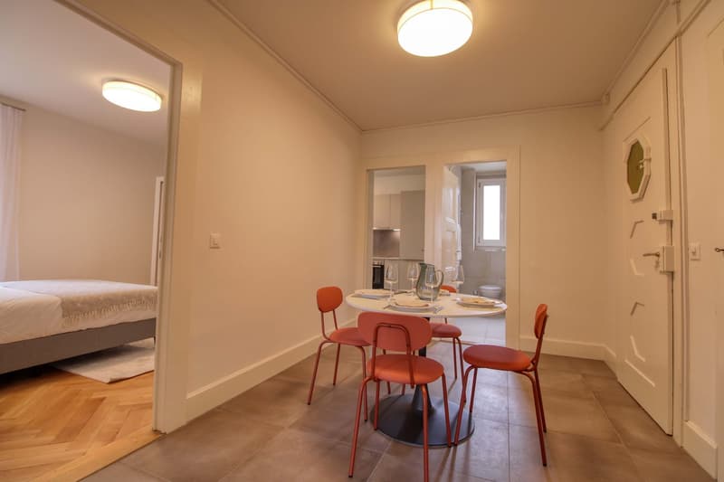Dining area with a round table, four chairs, and a small rug. A bed is visible in the adjacent room.
