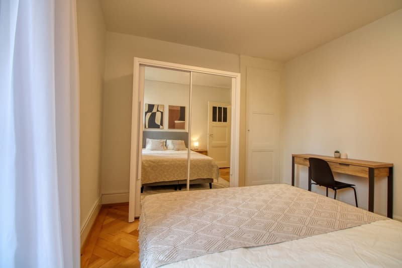 Bedroom with bed, desk, chair, mirror, and nightstand with lamp