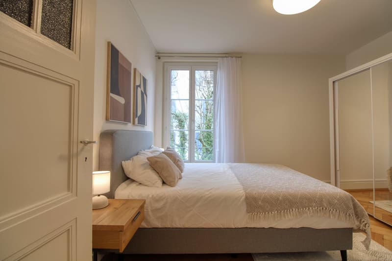 Bedroom with a double bed, nightstand, lamp, and large window with curtains. The room has a modern and clean design with white walls and wooden floors.
