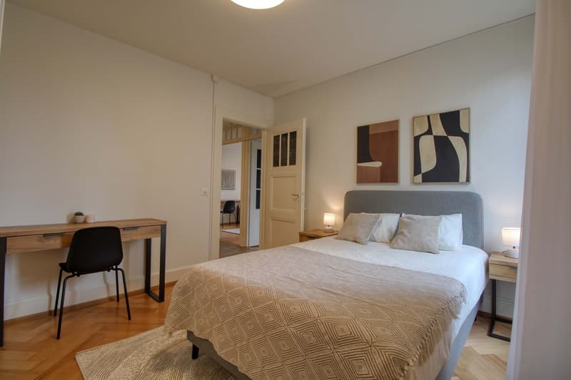 Bedroom with bed, desk, chair, nightstands, lamps, rug, and artwork on the wall.