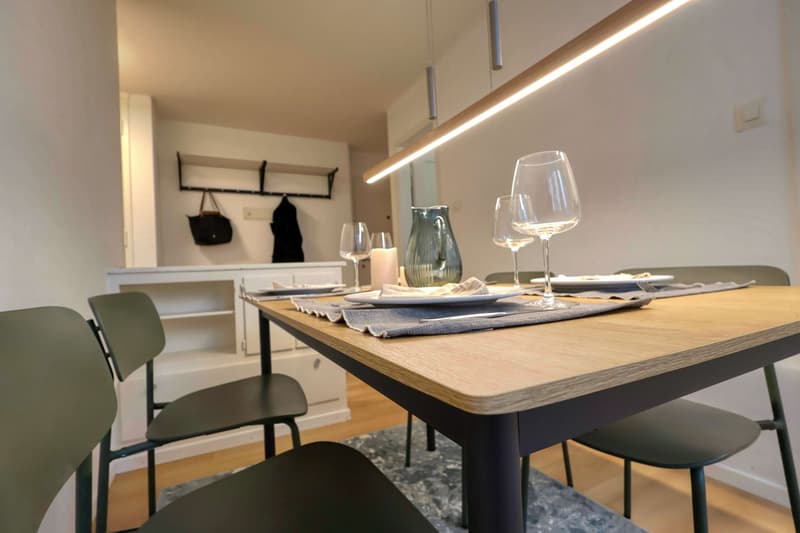 Dining room with a wooden table, four chairs, a white cabinet, a shelf with bags, and a lit ceiling lamp.