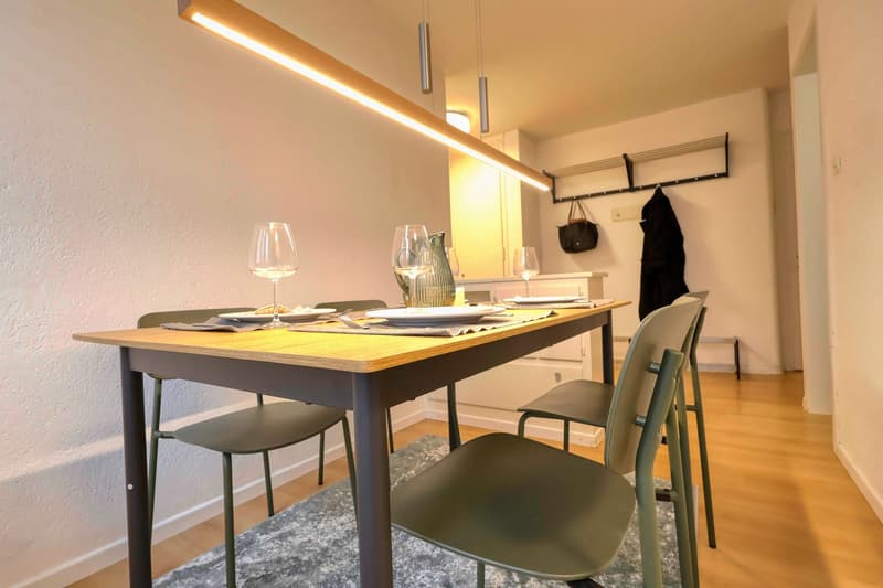 Dining room with a wooden table, four chairs, a rug, and a shelf with a handbag and a coat