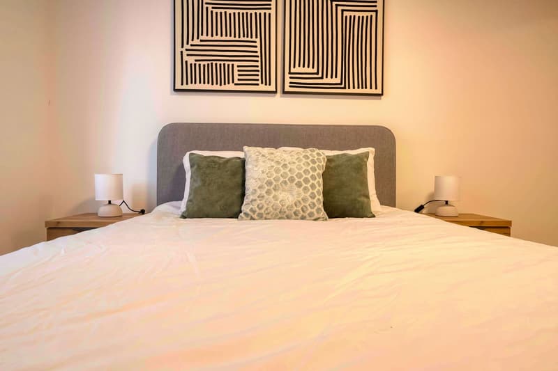 A neatly made bed with white sheets, two nightstands with lamps, and two abstract paintings on the wall.