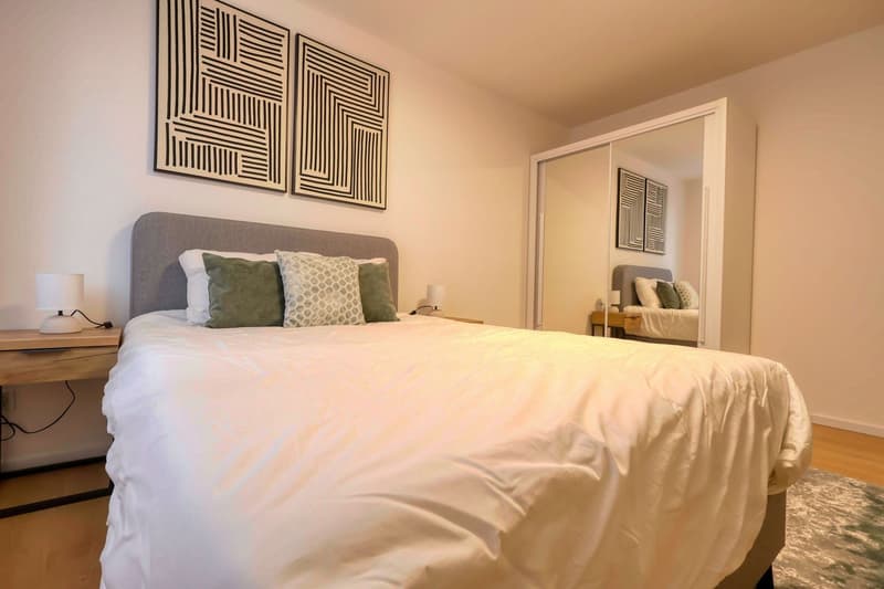 Bedroom with a bed, nightstands, lamps, and a mirror. The bed has a gray headboard and white bedding. The nightstands have lamps and a power outlet. The wall has two abstract paintings. The mirror reflects the room.