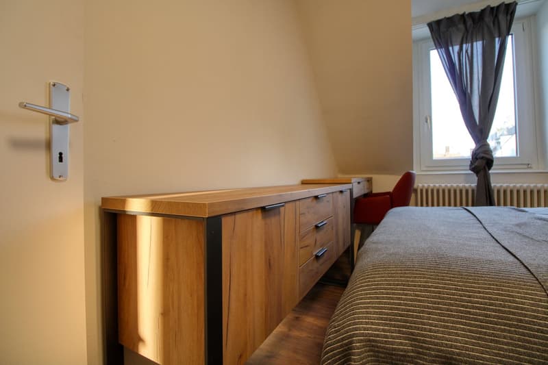 A bedroom with a bed, a wooden cabinet, and a chair beside the window