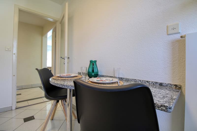 The image shows a kitchen and dining area with a round granite-topped table, two black chairs, a green vase, and a set of plates and glasses. The room has white walls and tile flooring, and there is a doorway visible in the background.