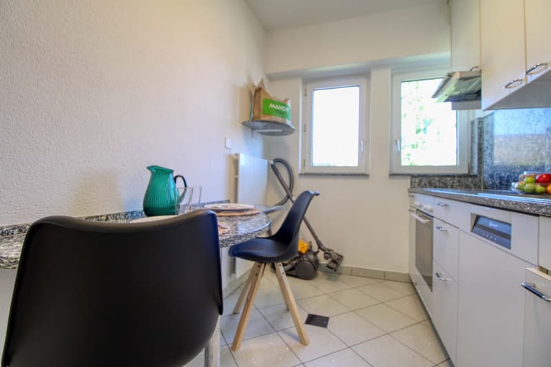 Compact kitchen with white walls, tile flooring, granite countertops, and modern appliances including a dishwasher. There is a small dining area with a table and chairs. The kitchen has ample storage space and natural light from the windows.