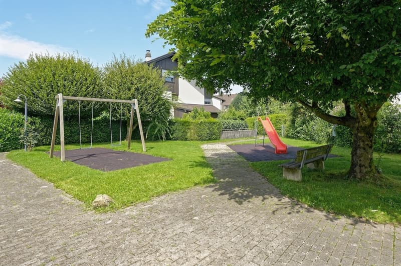 playground area with swings and slide, a bench, green lawn, a tree, and paved walkways