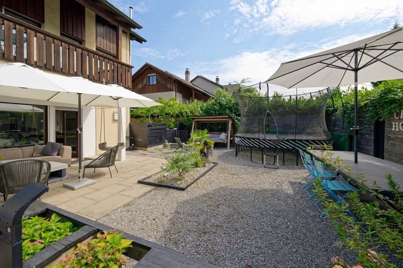 large gravel area, two white umbrellas, a few chairs, a table, plants, trees, trampoline, garden furniture