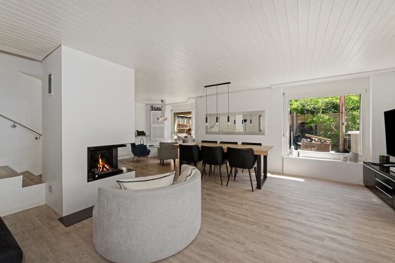 wooden floor, fireplace, open space with dining area, glass doors leading to garden, black chairs and table, grey sofa with pillows, TV