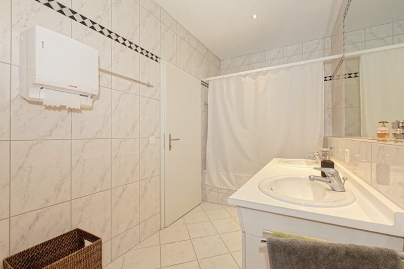 White tiled walls, white tiled floor, white sink, faucet, a mirror, paper towel dispenser, shower curtain