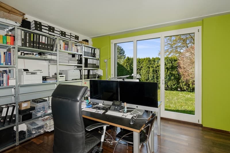 home office with black chair, desk with two computer monitors, shelves with books and files, green wall, sliding doors with grass view