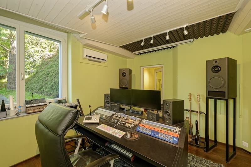 Professional recording studio, featuring black leather chair, control desk with multiple electronic devices, large monitor, speakers, air conditioning, and guitars on a stand.
