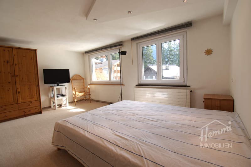 Bedroom with bed, wooden cabinet, TV, chair, radiator, windows, carpeted floor