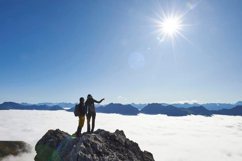 Two people standing on a mountain peak, pointing at the view, with a bright sun and fog below