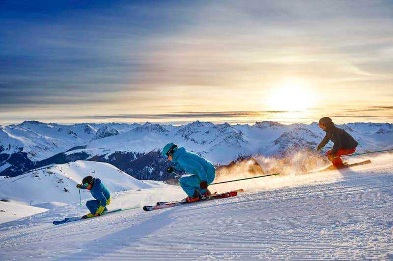 Three skiers on a snowy mountain slope, with a sunset in the background.