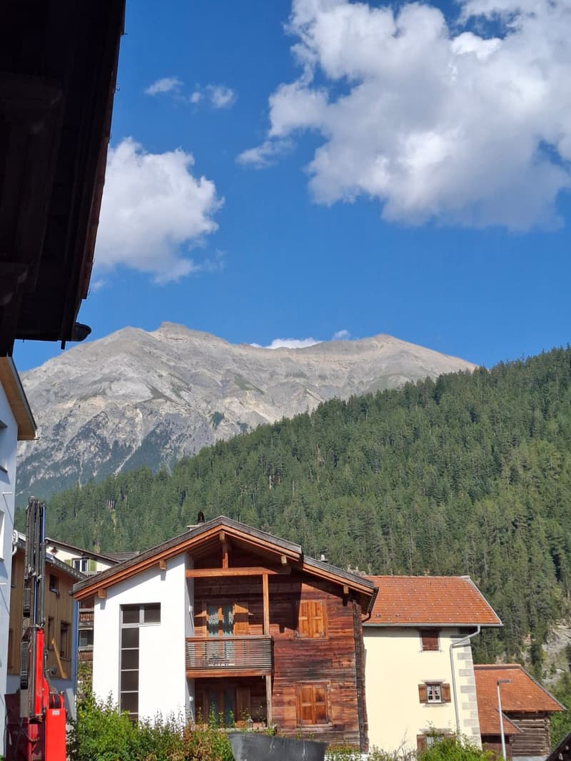 Mountain-style wooden and white house, balcony, lush trees, sunny day, blue sky, mountains in the background
