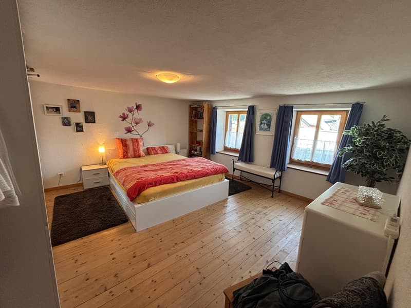 central bed, wooden floors, shelves against the wall, lamp on the nightstand, windows with blue curtains, rug, potted plant, white walls, pictures on the wall