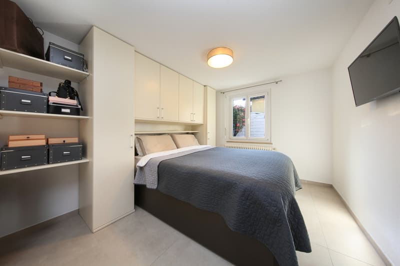 Spacious bedroom with built-in wardrobe, queen-size bed, and storage boxes on shelves. The room has a clean, modern design with white walls and a tiled floor. A small window provides natural light to the space.