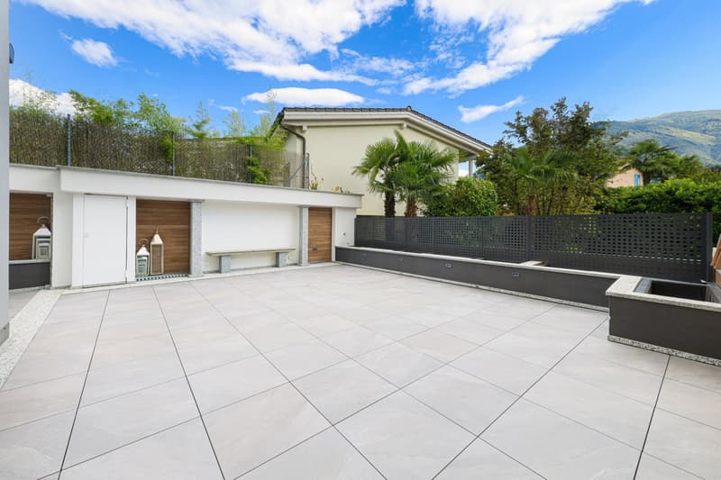 Patio with tiled floor, wooden fence, two wooden entrances, small benches, large greenery backdrop