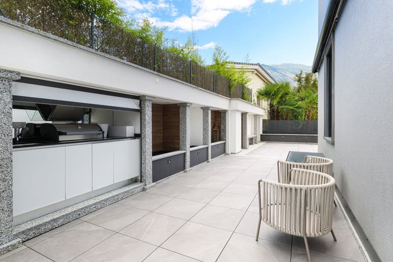 outdoor dining area, tile flooring, outdoor grill, two chairs, green hedge, distant building