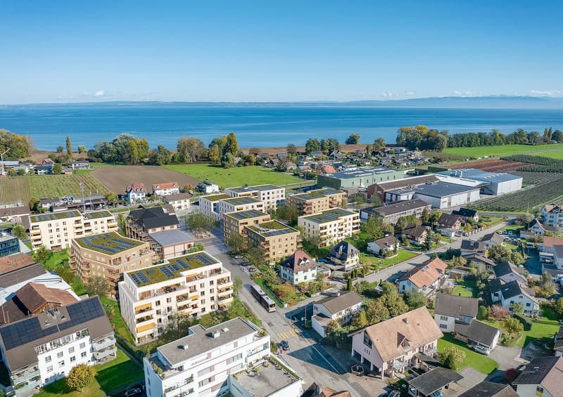This image shows an aerial view of a town or city located near a large body of water, likely a lake or sea. The town consists of a mix of residential and commercial buildings, with some multi-story apartment buildings and smaller houses visible. There are
