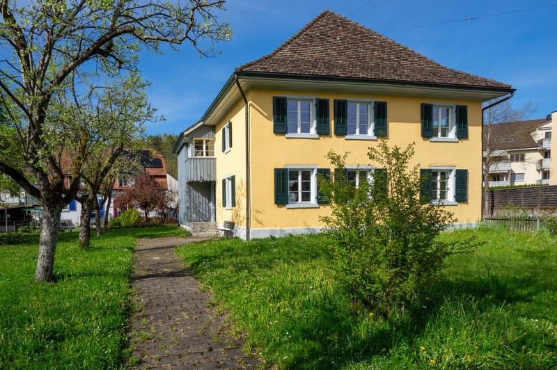 2 story house, yellow exterior, brown roof, green shutters, brick pathway, garden, trees