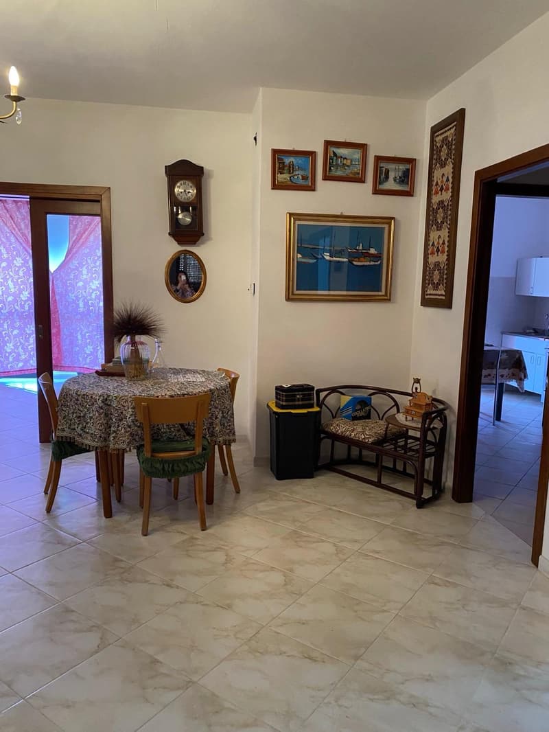 The image shows a cozy dining room with a round table, chairs, and various decorative elements. The room features a fireplace, wall art, and a cabinet with decorative items. The floor is tiled with a marble-like pattern, adding to the overall warm and inv
