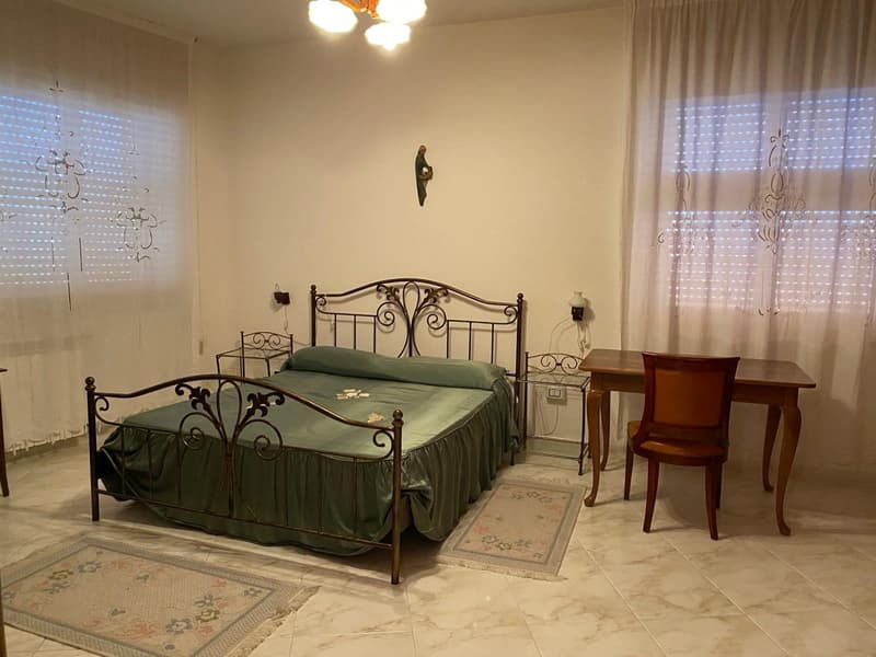 The image shows a bedroom with a metal bed frame with an ornate, scrolled design. The bed is made with a green bedspread. There is a wooden desk and chair in the room, as well as a decorative wall hanging above the bed. The room has tile flooring and curt
