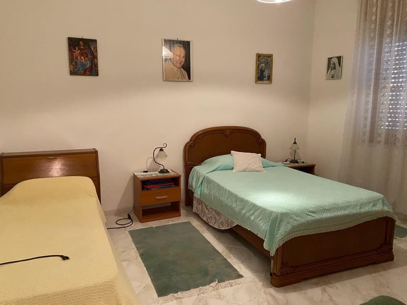 The image shows a bedroom with a wooden bed frame, a nightstand, and various religious artwork on the walls. The room has a tile floor and a curtain-covered window.