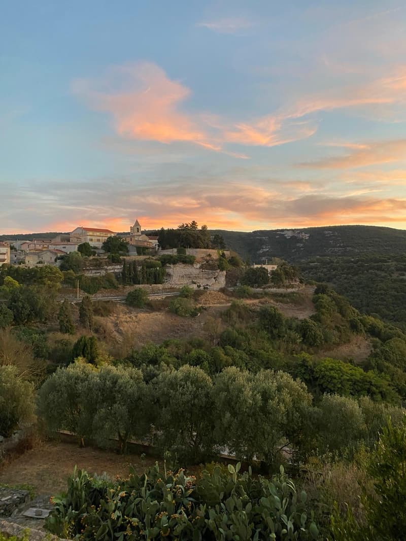 Scenic view of a hillside town with a church steeple, surrounded by lush greenery and a colorful sunset sky.