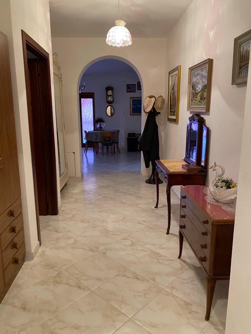 The image shows a spacious hallway with a marble tile floor. The hallway leads to a dining room area visible through an arched doorway. The hallway features decorative elements such as framed artwork, a vintage dresser, and a chandelier lighting fixture.