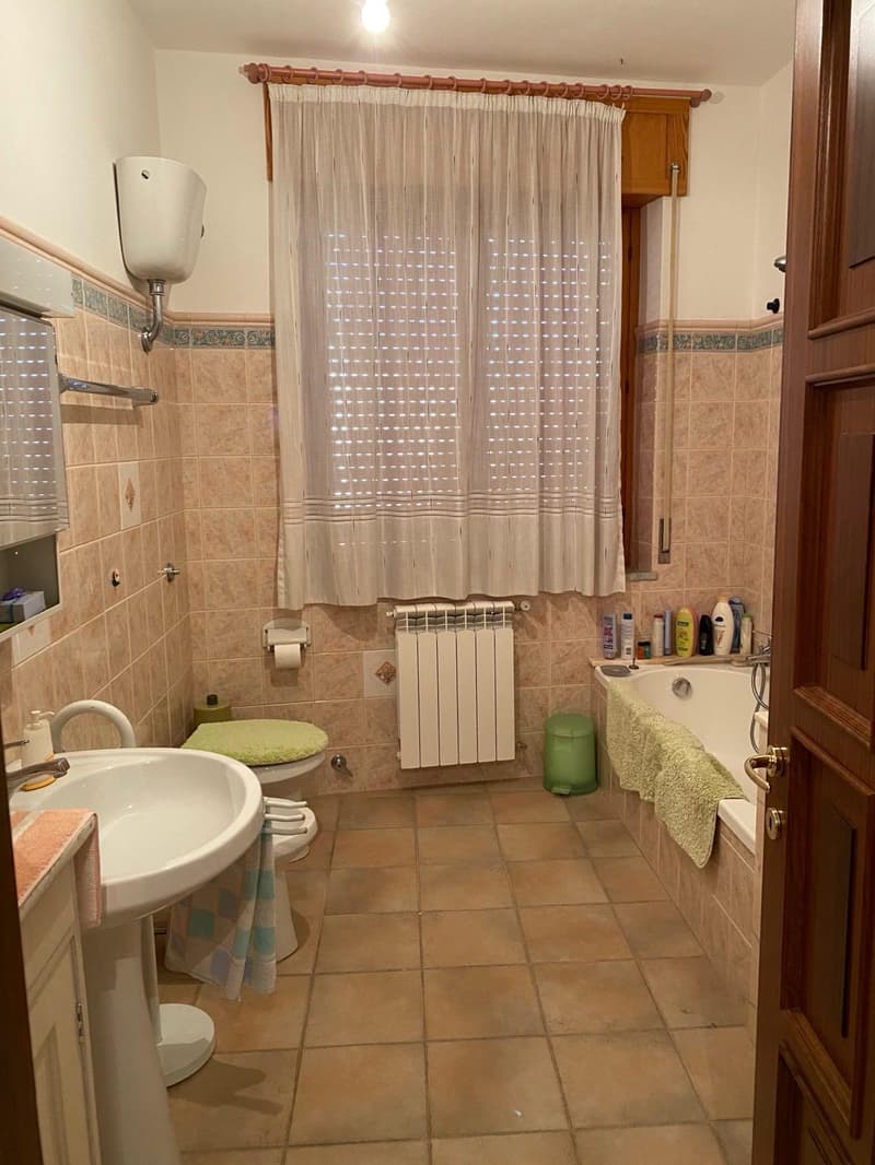 Bathroom with tile flooring, white toilet, sink, bathtub, curtain-covered window, and a radiator heater. There are various toiletries and personal items visible.