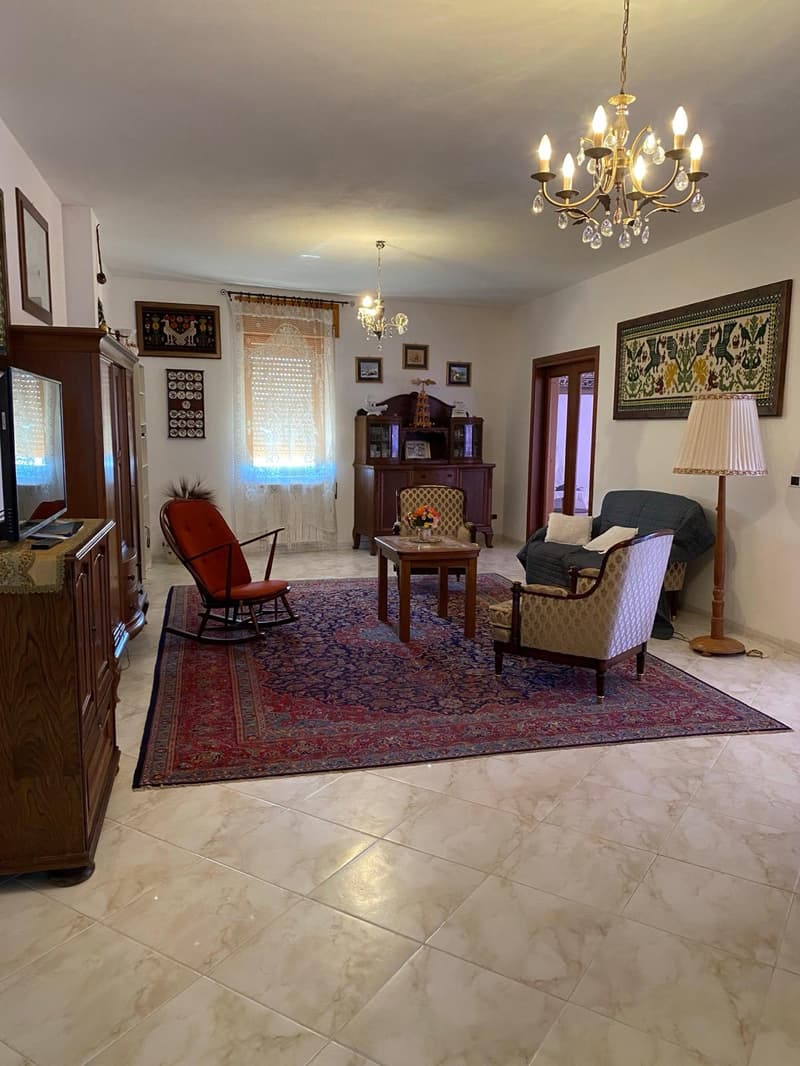Spacious living room with hardwood floors, large windows, a chandelier, a fireplace, and traditional furniture including a rocking chair, sofa, and coffee table. The room is decorated with framed artwork and other decorative elements.