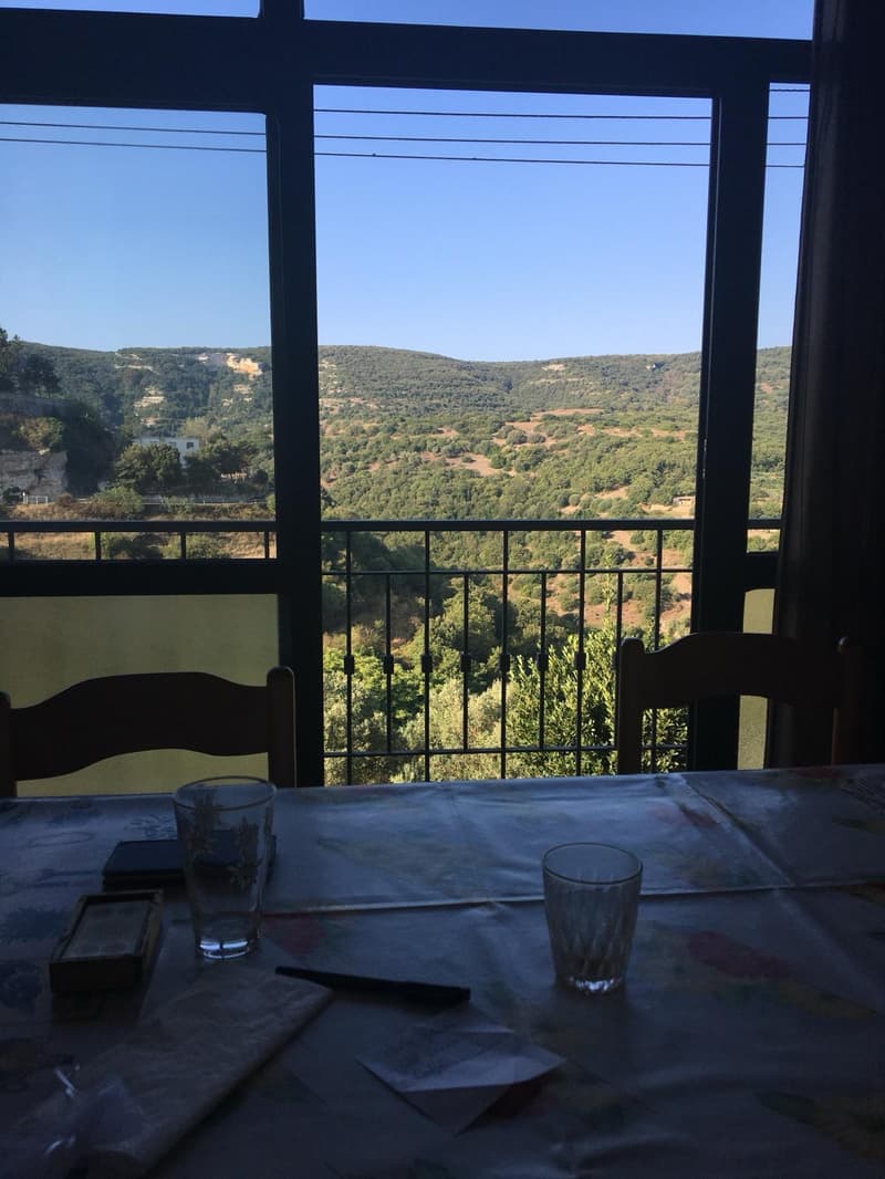 The image shows a balcony or terrace overlooking a scenic landscape. The view features rolling hills covered in lush greenery, with a clear blue sky in the background. The balcony is furnished with a table and chairs, and there are some personal items suc